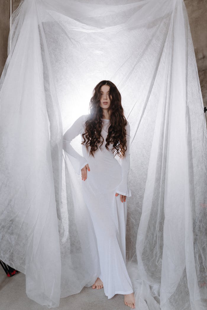 A woman in a long white gown stands between draped sheer fabric, creating a serene atmosphere.