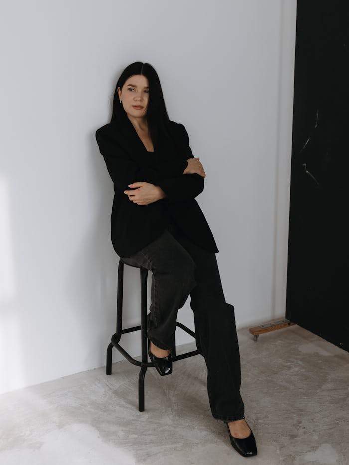 Confident woman sitting in stylish black clothes on a chair against a minimalist background.